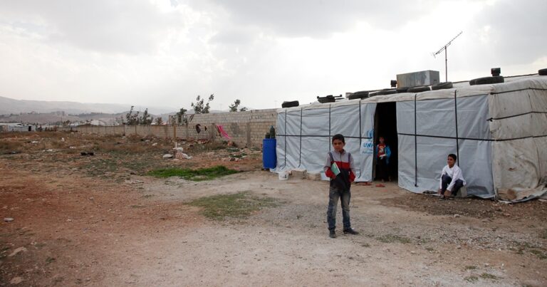 A young child stands outside a temporary shelter in a barren landscape, with another person visible in the doorway. The sky is overcast, indicating a potentially rainy day.