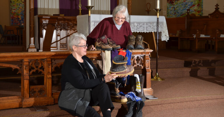 Two women are seated in a church setting, one holding a vintage hat while the other stands behind her, surrounded by various footwear displayed on a wooden altar. The scene captures a moment of sharing or discussion related to the items, emphasizing community and heritage.