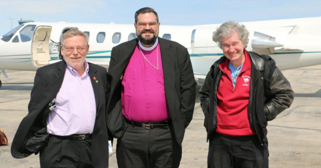 Three men standing together in front of a private jet on a sunny day, wearing formal and casual attire. The man in the center is dressed in a clerical collar and a purple vestment, flanked by two men in suits and casual jackets.