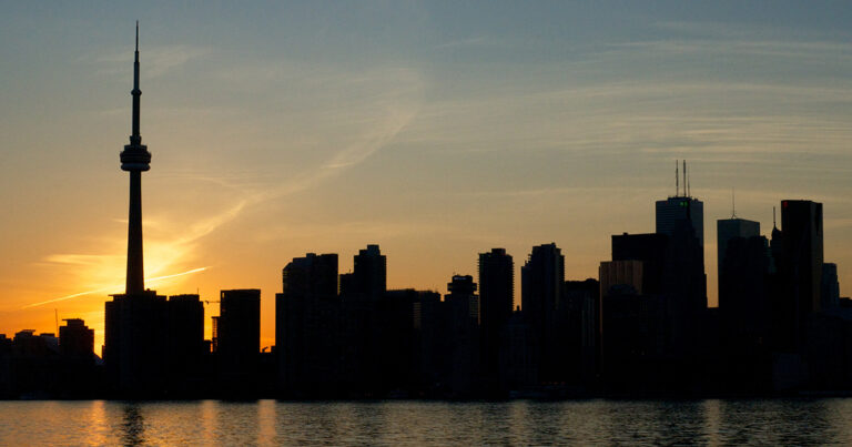 Silhouette of a city skyline at sunset, with buildings outlined against a colorful sky reflected in the water below.