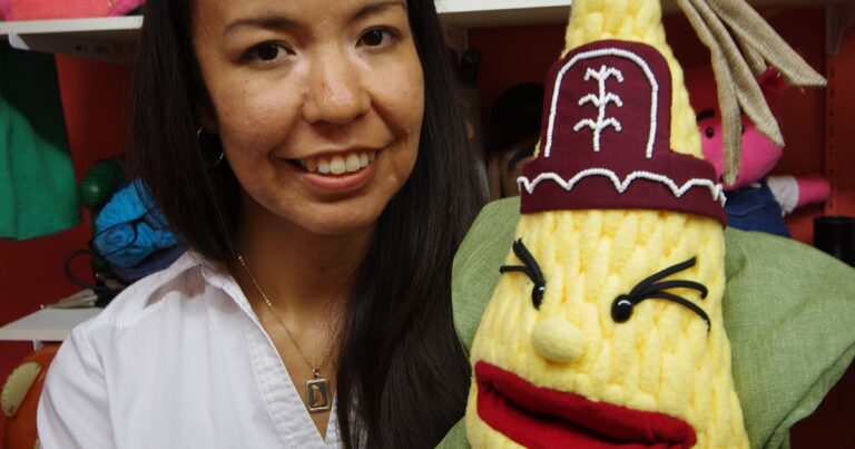 A woman smiles while holding a colorful puppet resembling a corn character, featuring a hat and expressive facial features.