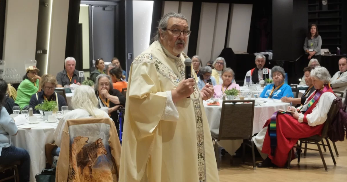 Archbishop in pale yellow chasuble speaks to a room of people seated at round tables.