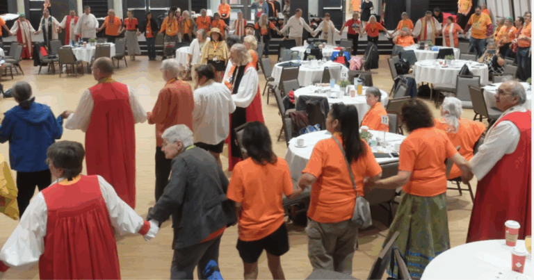 A circle of people in orange shirts and religious robes hold hands in prayer.