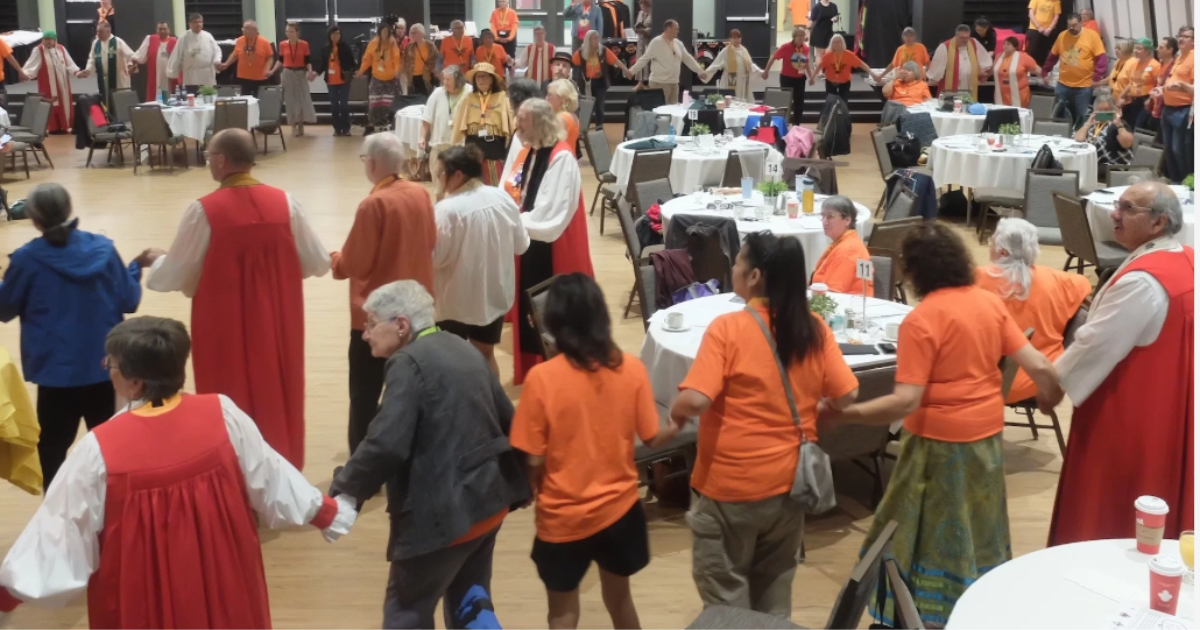 A circle of people in orange shirts and religious robes hold hands in prayer.