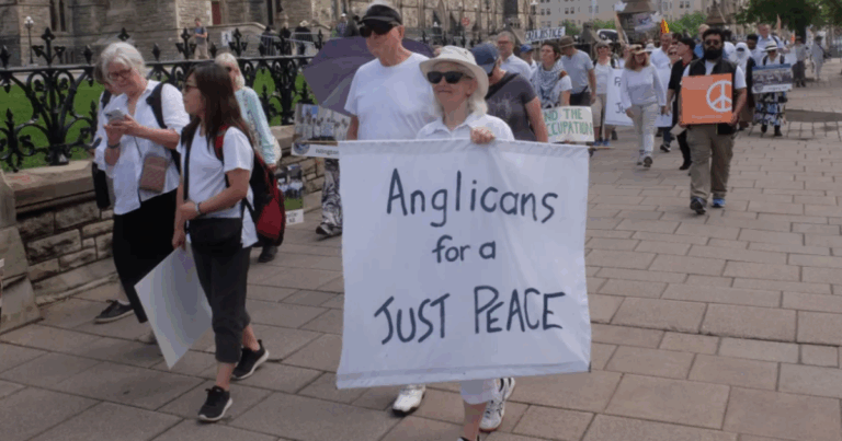A participant in a peaceful demonstration holds a sign reading "Anglicans for a Just Peace," surrounded by fellow marchers wearing white. The setting appears to be an outdoor public space.