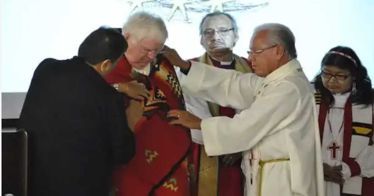 Archbishop Michael Peets is wrapped in a red blanket adorned with traditional Indigenous patterns during a ceremonial moment led by Indigenous clergy and community members