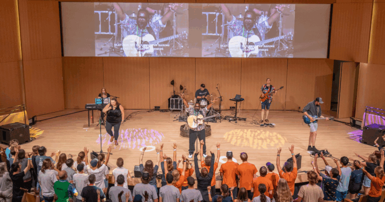 A lively music performance taking place on stage with a band, featuring musicians playing instruments and vocalists engaging with an enthusiastic audience. Large screens display visuals of the performance, enhancing the concert atmosphere.