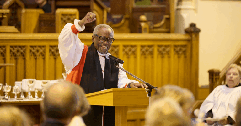 A smiling speaker in a clerical robe raises his fist while addressing an audience in a church setting, with wooden architecture in the background.