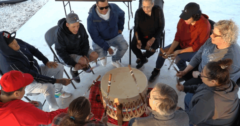 Group of people sitting in a circle, playing a large drum together, with a focus on community engagement and cultural expression.