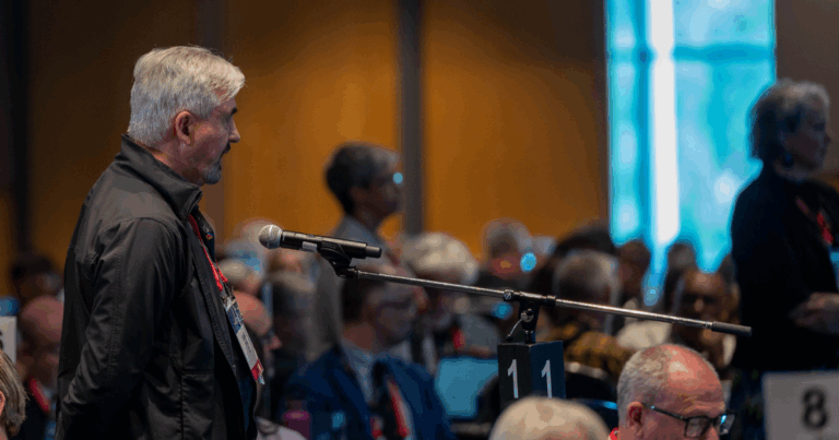 A man with gray hair and a black jacket speaks into a microphone at a conference. He stands among a seated audience in a dimly lit room.