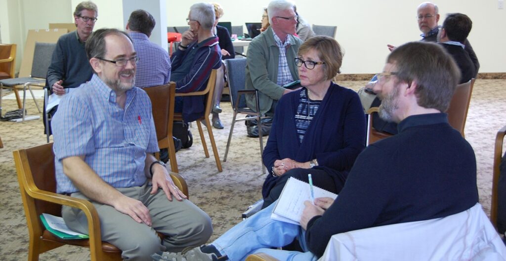 Group discussion among older adults in a community center, with three individuals engaged in conversation while others are seated in the background, participating in various activities.