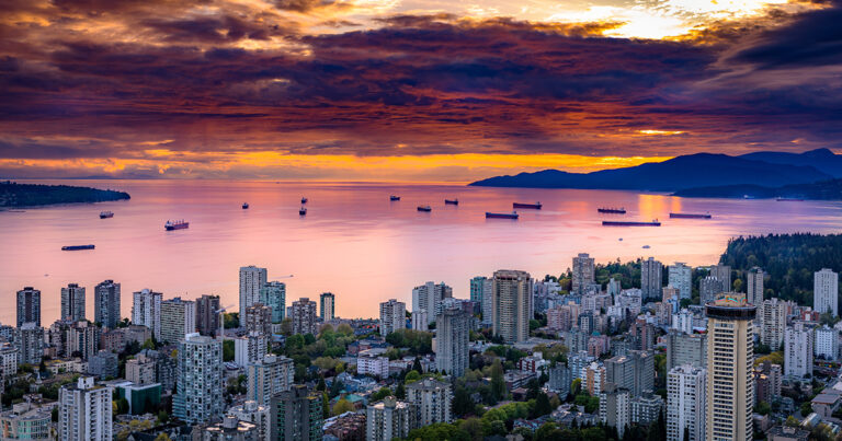 Aerial view of Vancouver at sunset, showcasing a skyline of high-rise buildings with the ocean and ships in the background, under a colorful sky.