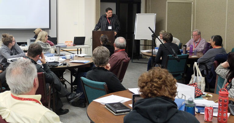 A speaker presenting at a conference in a meeting room, with an audience seated at tables listening attentively.