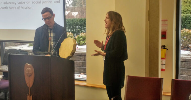 A woman in a black dress stands at a podium, gesturing while speaking to an audience. A man is positioned next to her, holding a circular award or plaque. The setting appears to be a formal event or presentation.
