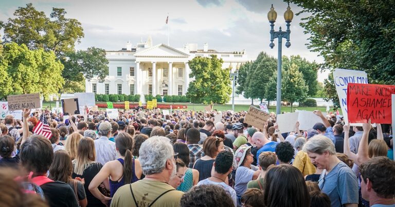 Crowd of protesters gathered in front of the White House, holding signs and demonstrating during a rally. The scene captures a diverse group of individuals engaged in activism against a backdrop of the iconic building.