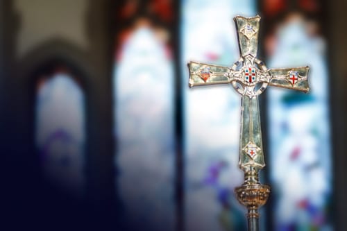 The Primatial Cross of the Anglican Church of Canada, an ornate silver processional cross, stands before softly blurred stained-glass windows inside a church.