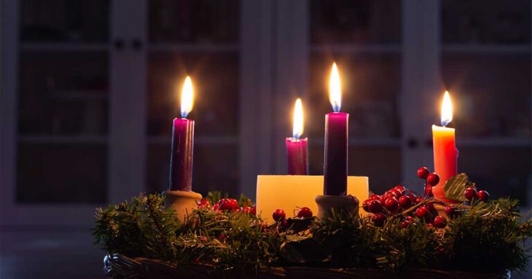 Three lit candles, two purple and one white, surrounded by greenery and red berries, creating a warm and festive ambiance.