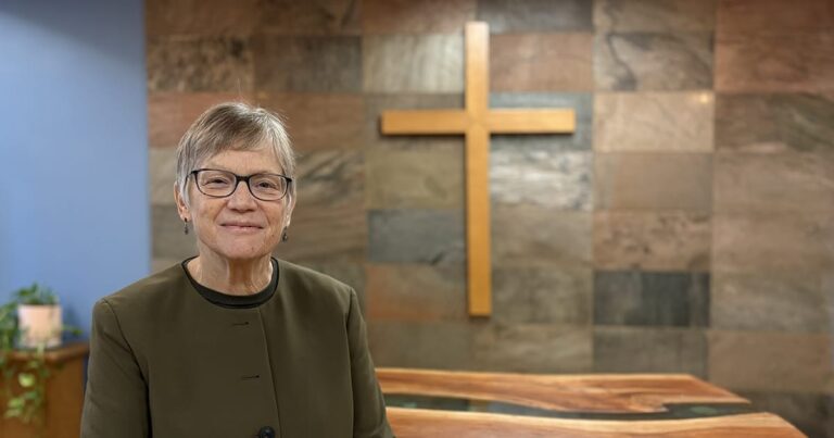 A middle-aged woman with short gray hair and glasses, wearing a dark green blazer, smiling in front of a wooden cross and a stone wall background.