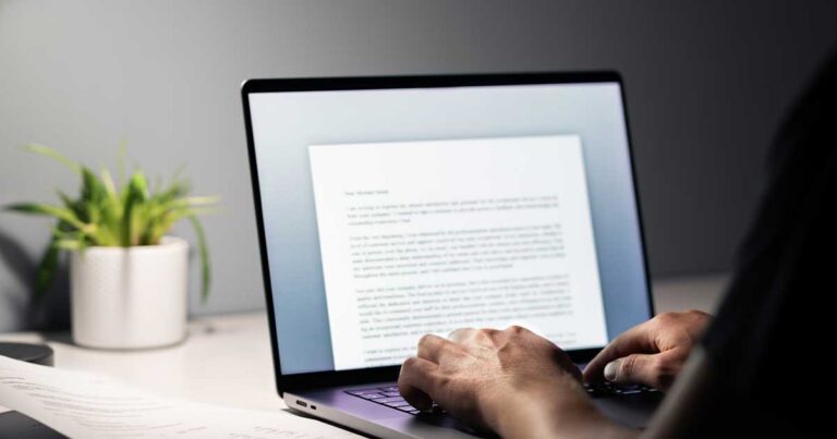 A person types on a laptop at a desk, with a document open on the screen. The scene is softly lit from the side, and a small green plant in a white pot sits nearby.