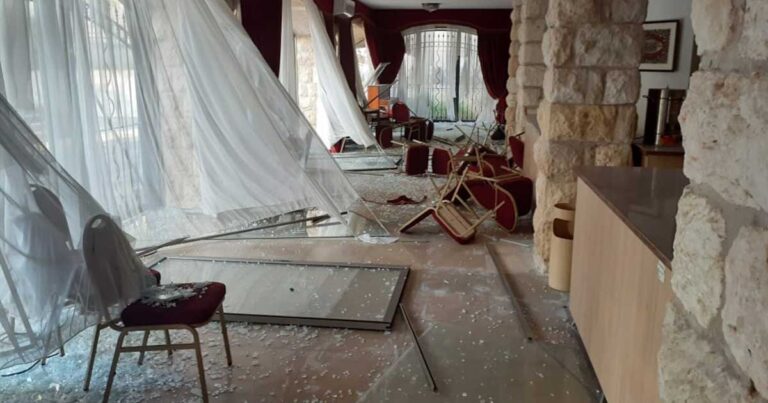 Interior of a damaged room with broken furniture, shattered glass, and torn curtains, indicating signs of destruction and disarray.