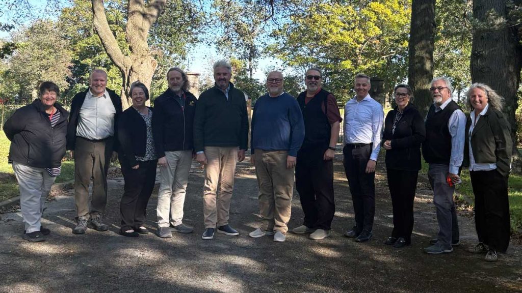 Eleven people stand together outdoors beneath large trees, smiling for a group photo on a sunny day.