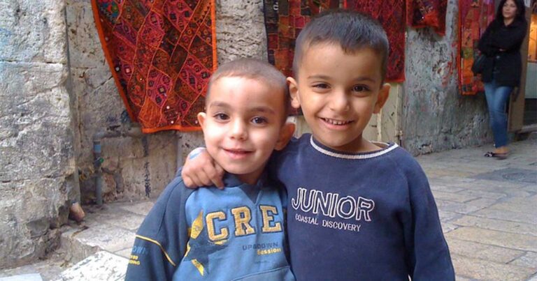 Two young boys smiling and posing together, one with short hair wearing a blue sweatshirt with "CREW" printed on it, and the other in a navy blue "JUNIOR" sweatshirt, standing against a textured background.