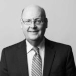 A middle-aged man in a dark pinstripe suit, striped tie, and glasses, smiling slightly while facing the camera against a plain light background.
