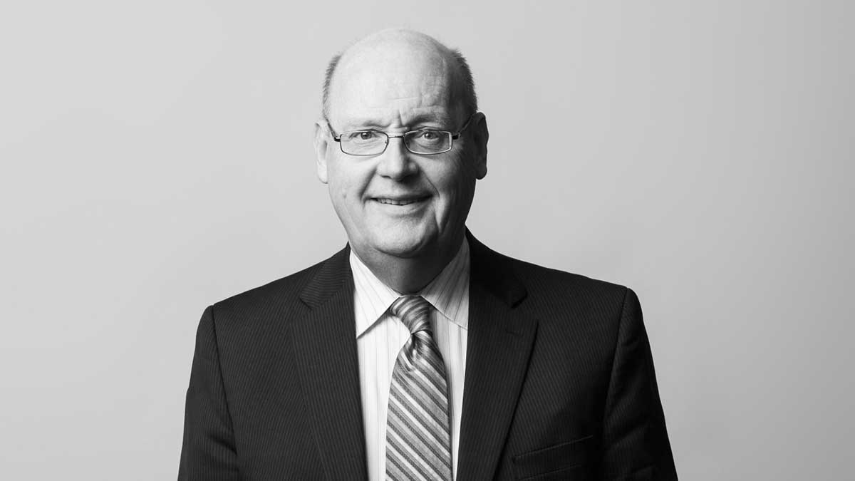 A middle-aged man in a dark pinstripe suit, striped tie, and glasses, smiling slightly while facing the camera against a plain light background.