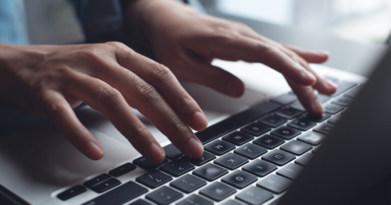A person types on a laptop at a desk, with a document open on the screen. The scene is softly lit from the side, and a small green plant in a white pot sits nearby.