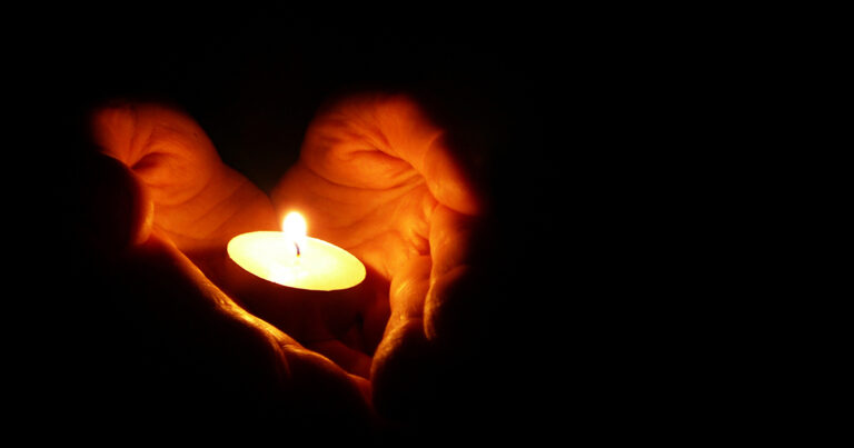 A pair of hands gently cradling a lit candle against a dark background, symbolizing warmth and tranquility.