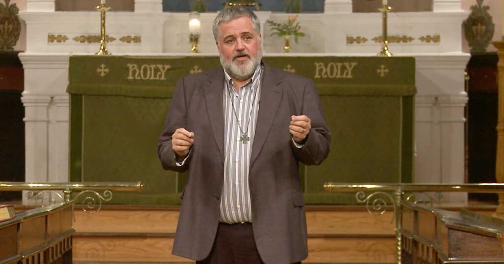 A man in a suit gestures while speaking on stage in a church setting, with a backdrop that includes the word "HOLY."