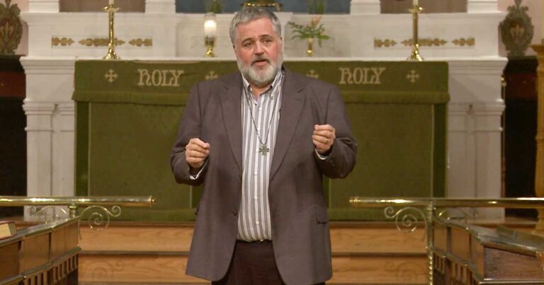 A man in a suit gestures while speaking on stage in a church setting, with a backdrop that includes the word "HOLY."
