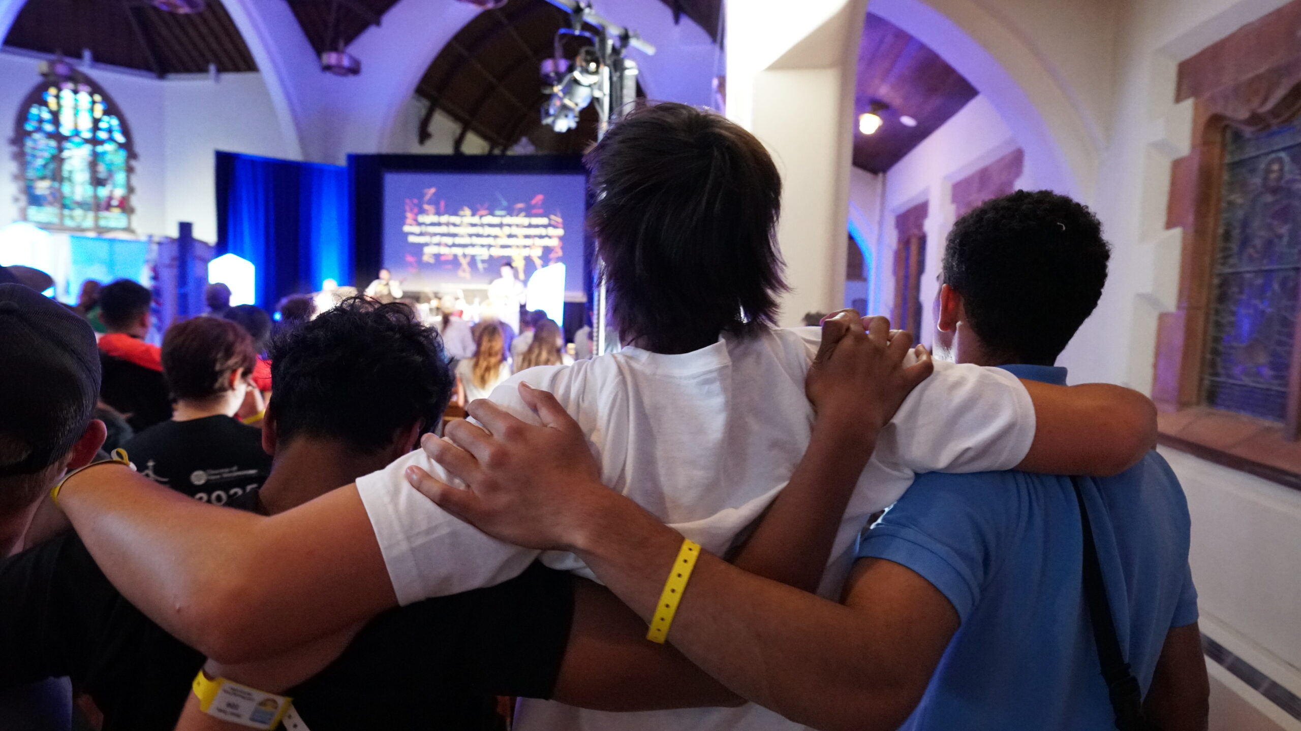 A group of people with their arms around each other, watching a presentation on stage in a well-lit venue with an audience.