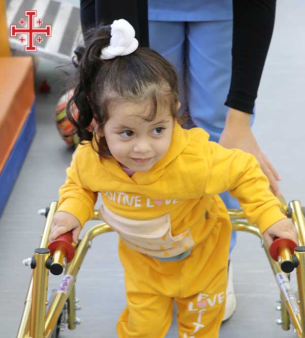 Young child in a yellow outfit using a pediatric walker during a therapy session, supported by an adult standing behind her; the Companions of Jerusalem logo—a red Jerusalem cross with small crosses in each quadrant—appears in the top-left corner.