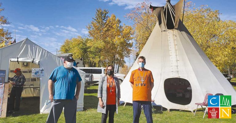Three individuals wearing face masks stand outdoors in front of a large tipi, surrounded by trees with autumn foliage. The person on the left wears a blue shirt, the middle person wears a gray coat, and the person on the right wears an orange shirt. The setting suggests a cultural or community event.