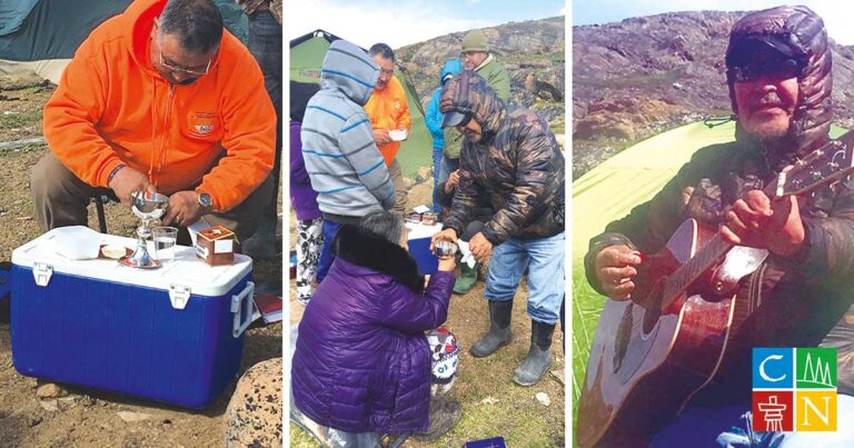 Group of people preparing food outdoors in a rugged landscape, with one person seated and others engaged in cooking activities, wearing warm clothing.