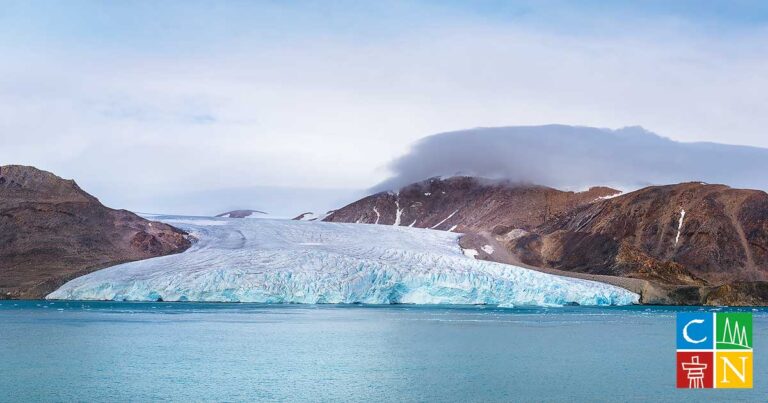 A panoramic view of a glacier melting into a calm body of water, surrounded by rugged mountains and a cloudy sky, illustrating the effects of climate change.