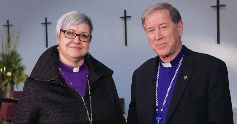 Two clergy members, one with short gray hair and glasses and the other with short hair, both wearing purple vestments, stand together in front of a wall with a cross.