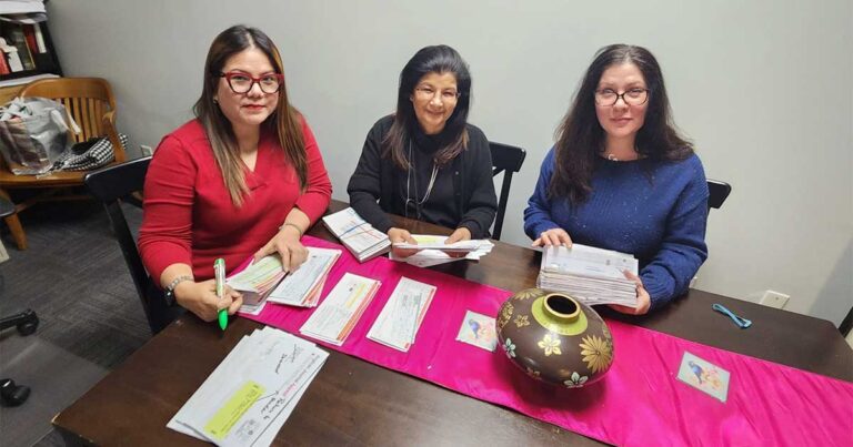 Three women engaged in a collaborative discussion at a table, surrounded by papers and a decorative pot, with a vibrant pink tablecloth.