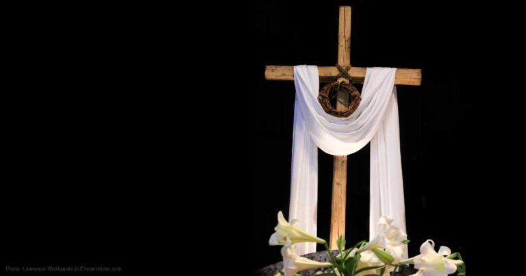 Wooden cross adorned with a white drape and surrounded by white lilies, set against a black background.
