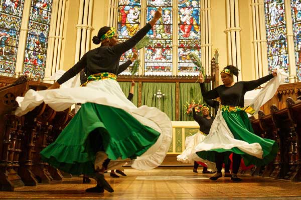 Liturgical dancers in flowing green, white, and black garments perform in front of stained glass windows inside a church sanctuary, their skirts sweeping outward as they dance in worship.