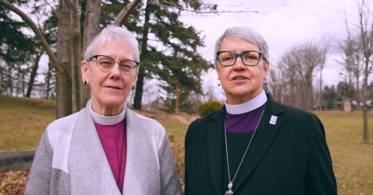 Two women standing outdoors, both wearing clerical attire. One woman has short gray hair and glasses, while the other has longer gray hair and is wearing a necklace with a pendant. They are positioned in front of trees and a grassy area, conveying a sense of community and leadership.