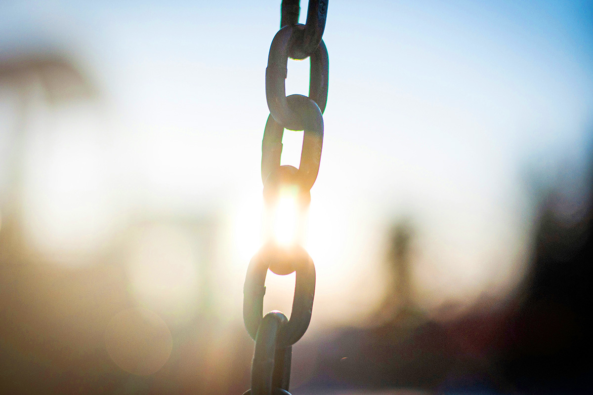 Close-up of a metal chain hanging vertically, backlit by sunlight that flares through the links against a softly blurred outdoor background.