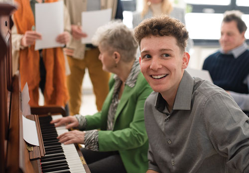 Young man smiling to the camera while woman plays piano sitting behind him. A Crowd listens in the background.