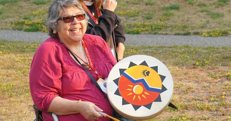 Smiling woman holding a decorative drum featuring a sun and mountain design, wearing glasses and a red shirt, outdoors during a community event.