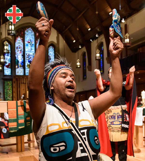 Indigenous dancer inside a church raising carved wooden rattles during a ceremonial moment, wearing regalia with Northwest Coast designs; other participants stand behind him in traditional clothing, with stained-glass windows visible in the background, and the Anglican Church of Canada crest in the top-left corner.