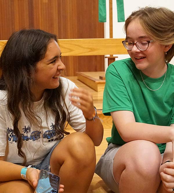 Two youth sit together and talk inside a church during a group gathering, with one wearing a white T-shirt and the other wearing glasses and a green shirt