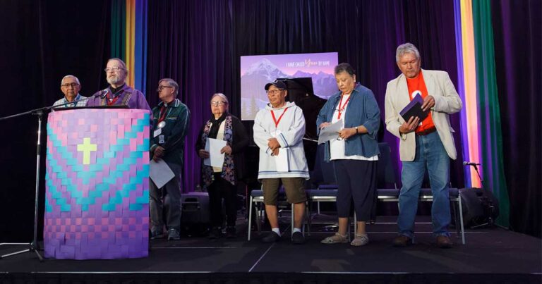A group of six individuals standing on stage during a ceremony, with a podium in the foreground and a colorful backdrop. They are holding papers and appear to be engaged in a presentation or speech.