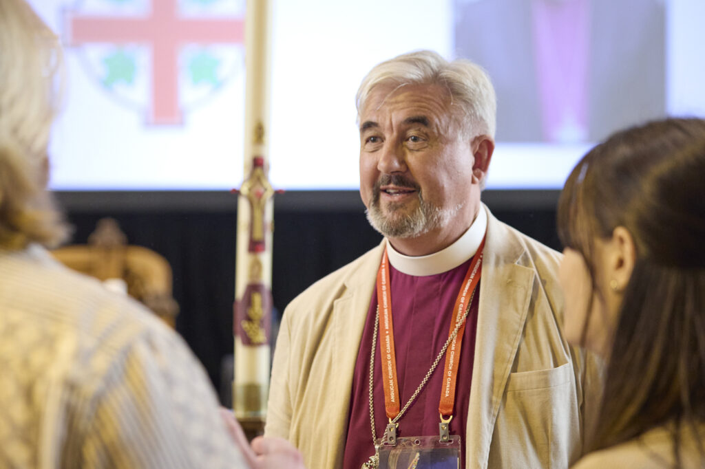 A smiling clergy member wearing a clerical collar and a vestment, engaging in conversation at an event, with a candle and a screen featuring a cross in the background.
