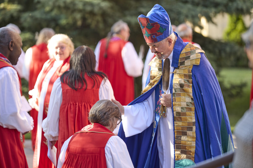 A group of individuals in colorful traditional attire participating in a ceremonial event outdoors, with one person in a blue robe and hat interacting with another.